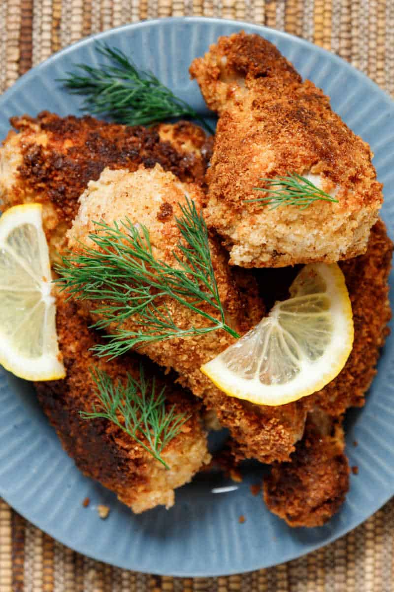 An image displaying a Keto Fried Chicken Recipe - crispy, golden-brown chicken pieces, fried to perfection and served on a white plate. The chicken's texture appears crunchy on the outside, yet remains succulent and juicy on the inside. On the side, you can see garnishes of fresh herbs, along with a dipping sauce, adding a vibrant color contrast to the golden fried chicken.
